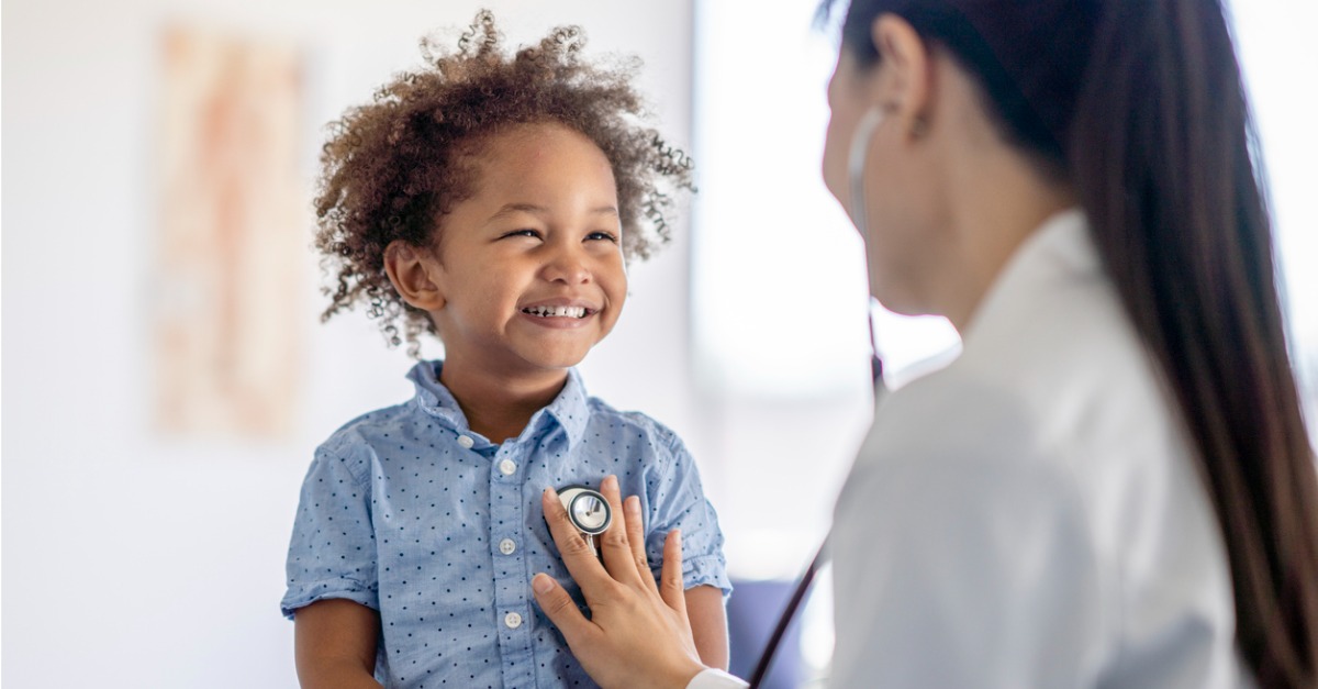 Photo showing a young boy smiling while a healthcare worker provides patient care, checking his heartbeat.