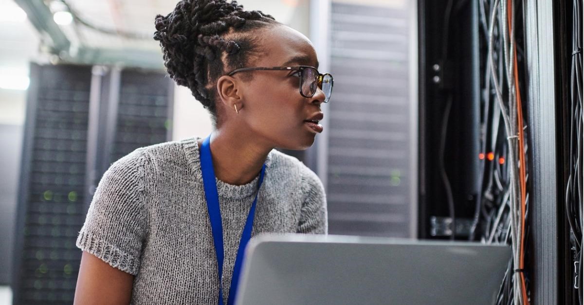 Photo of a tech worker holding a computer in a server room, addressing technical issues and resolving support tickets.